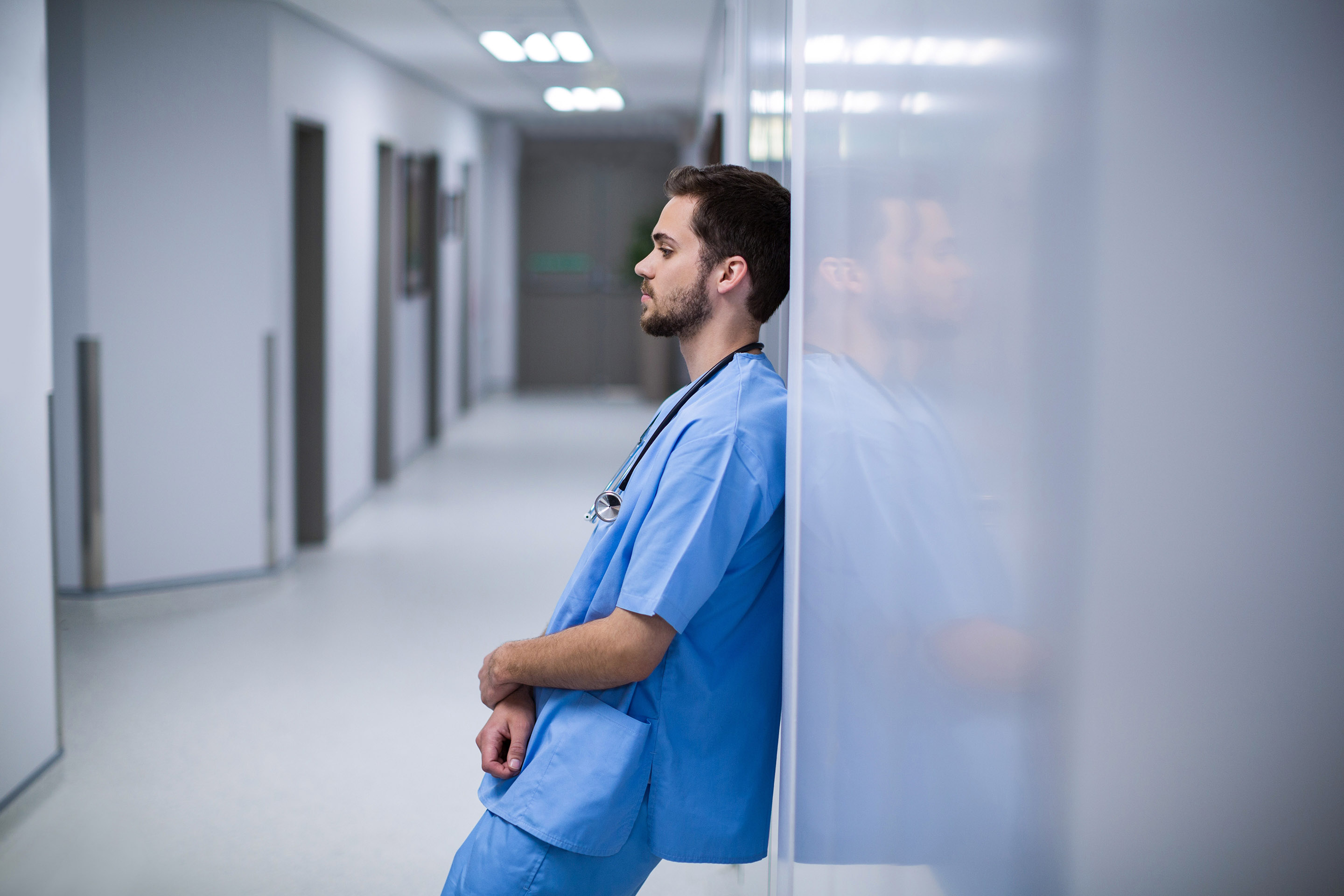 nurse in scrubs leaning in hallway, looking contemplative
